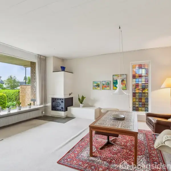 Stue med brunt lædersofaarrangement omkring et træbord. En rød orientalsk tæppe dækker gulvet. Store vinduer åbner til en velplejet have. Der står en lampe ved siden af sofaen.
