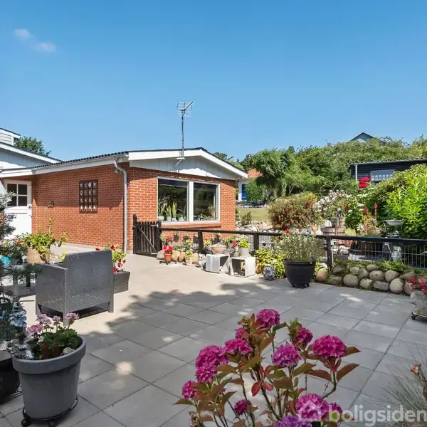 Rødt murstenshus med vindue stående i en have med blomster i krukker. Terrassen er flisebelagt med parasol, og det hele er omgivet af frodig vegetation under en klar blå himmel.