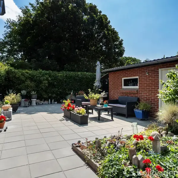 En solrig terrasse med grå fliser er omringet af farverige blomster i potter. En rød murstensbygning med vindue og dør danner baggrunden. Grønt hegn skaber en intim atmosfære.