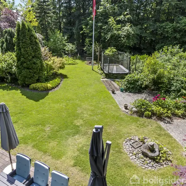 Grøn have med græsplæne omkranset af buske. En trampolin står i hjørnet på flisebelægning. I forgrunden står et terrassebord med stole og parasoller.