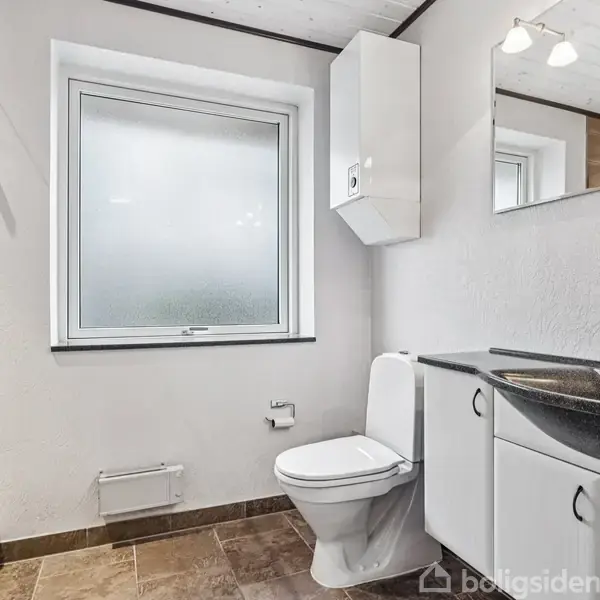 Toilet er placeret mellem et stort vindue og en sort vask med hvide skabe. Badeværelset har brune fliser, en brusekabine til venstre og hvidmalede vægge.