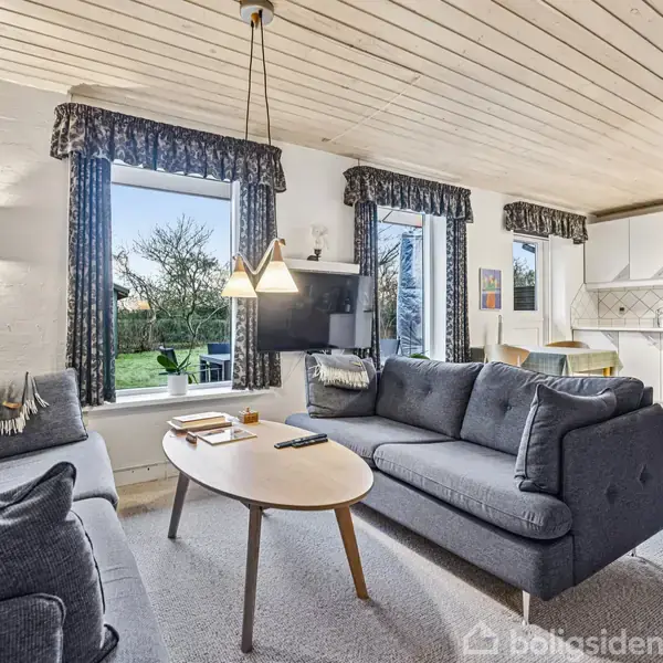 To grå sofaer står overfor hinanden med et ovalt sofabord imellem. Rummet har vinduer med udsigt til en have. Køkkenet er i baggrunden med spisebord og stole.