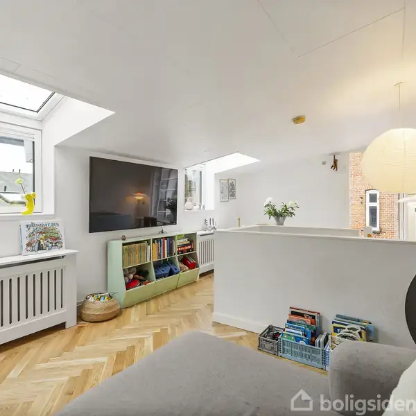 En hyggelig stue med sofa, fladskærms-TV på væggen, og en lav reol med bøger og legetøj. Ovenlysvinduer slipper naturligt lys ind og en lampe hænger fra loftet.