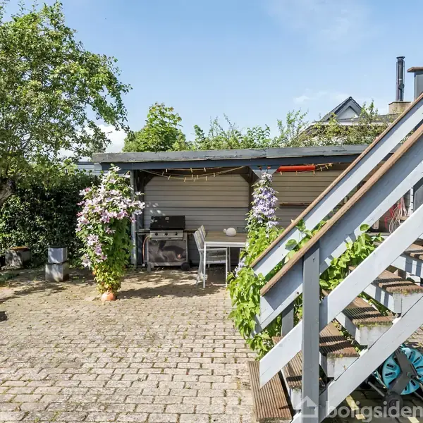 Trappe fører op til terrasse omgivet af farverige planter og blomster. I baggrunden står et skur med en grill. Sollys skinner på gårdhavens brostensbelægning.