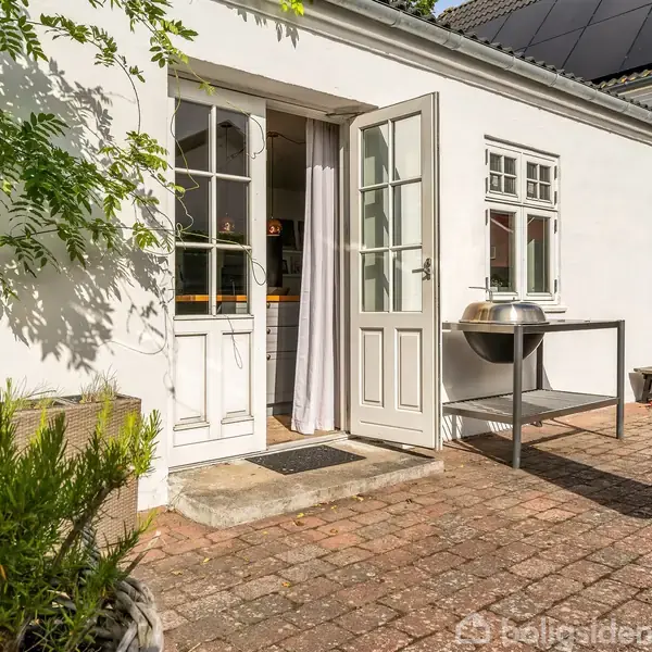 Dør står åben mod terrassen, flankeret af buske. Udekøkkenbord med grill står ved den hvide husmur. Solceller på taget. Terrassefliser dækker jorden. Ingen tekst fundet.