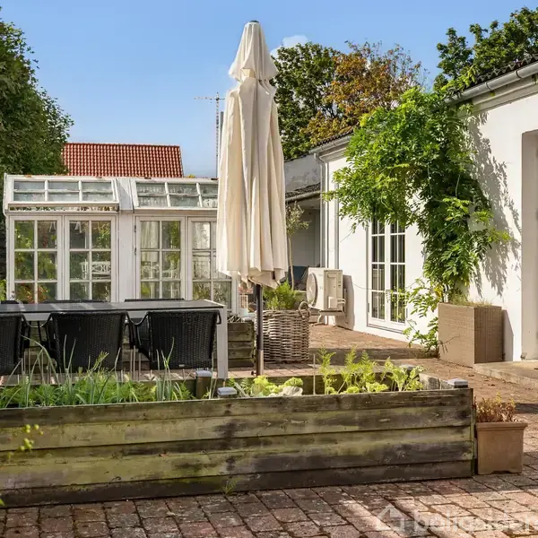 En lille gårdhave med et træplantebed fyldt med blomster og planter. Midt i står en parasol. Til venstre ses et drivhus, og til højre er en hvid bygning.