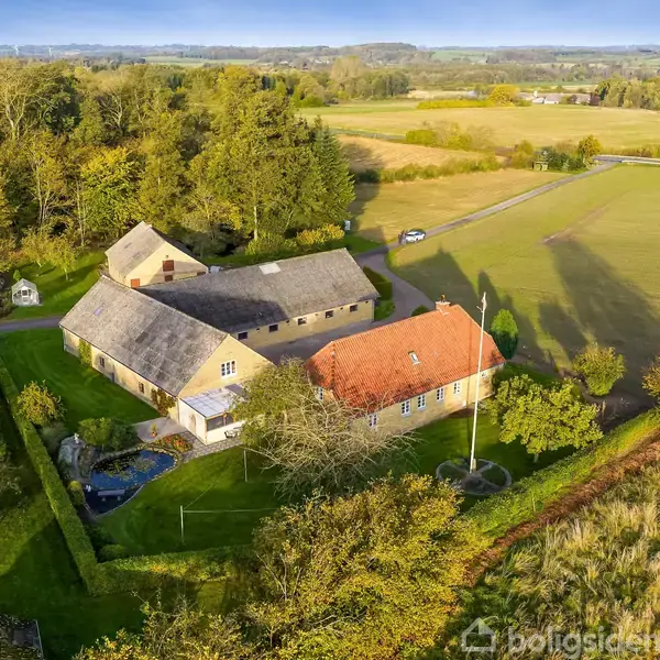 En gård med flere sammenhængende bygninger ligger omgivet af grønne marker og træer under en klar blå himmel. En grusvej fører til ejendommen fra en åben markvej.