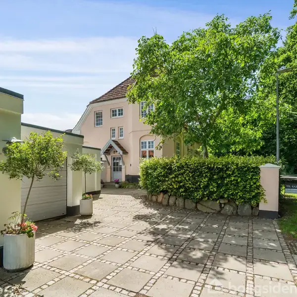 Et lyserødt hus med rødt tag står ved siden af en flisebelagt indkørsel. To træer og buske omgiver området, med en skovagtig baggrund i solskin.