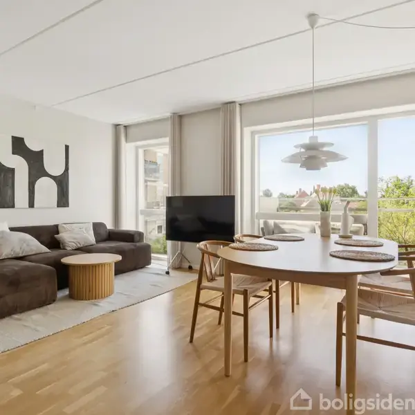 En moderne stue med brun sofa og lyst træbord. Store vinduer med udsigt til grønt område udenfor. Lyse trægulve og minimalistisk indretning med vægkunst og belysning.