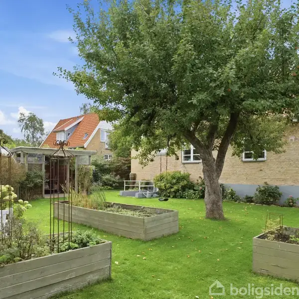Et stort træ kaster skygge over en grøn have. Forhøjede bede fyldt med planter er opstillet omkring træet. En lys stenbygning står i baggrunden, omgivet af buske og en terrasse.