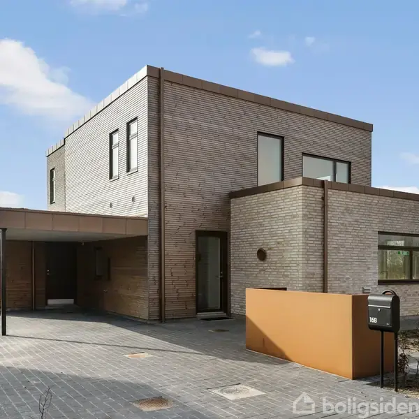 Moderne murstenshus med fladt tag står i en åben, landlig have. Indkørsel har en integreret carport og postkassenummeret er 142. Himlen er blå med lette skyer.