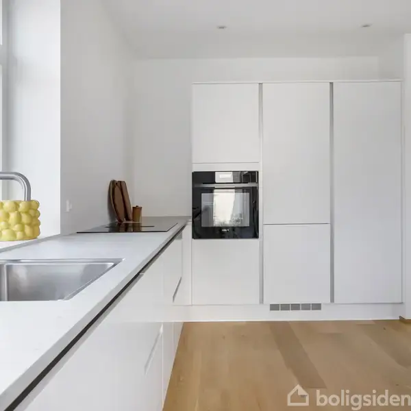 Et moderne køkken har hvide skabe og en indbygget ovn, ligger i et rum med trægulv og et stort vindue. Ved vinduet står en håndsæbepumpe og en skål citroner.