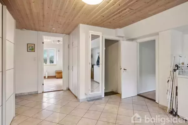 Entré med flisegulv og træbeklædt loft, indeholdende skabe med spejl. Døre fører til andre rum, hvorigennem et værelse med vinduer og lyst interiør kan anes. Skistave står i et hjørne.