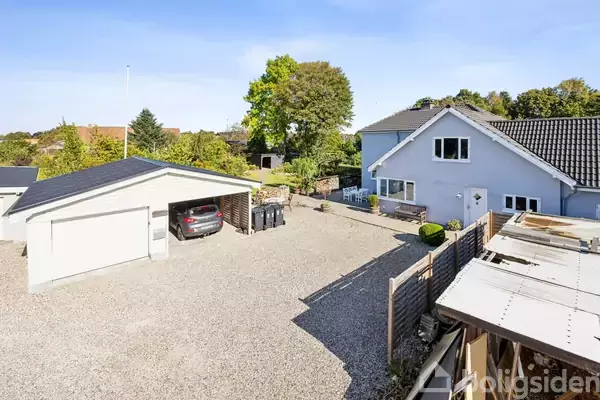 En grå bygning med skråt tag står ved siden af en carport med en bil parkeret. Omkring bygningerne ses grusbelagt gårdsplads, træhegn og frodige træer i baggrunden.