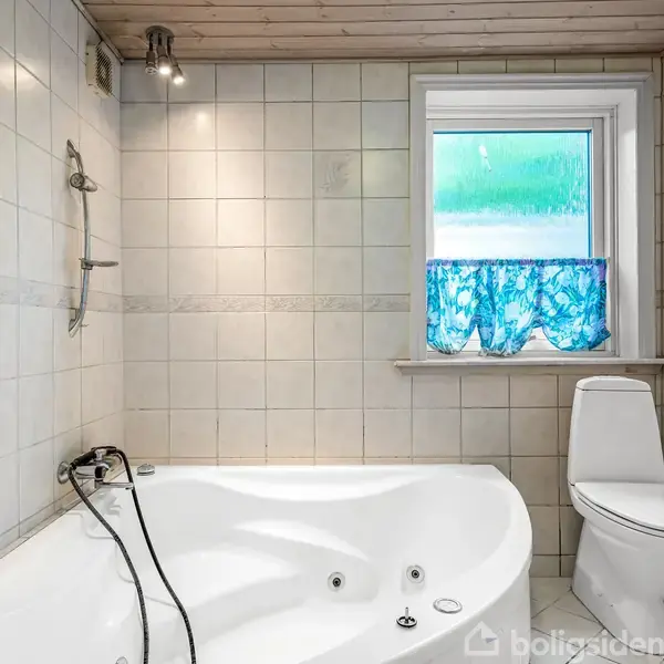 Et hjørnebadekar med vandhaner står i et flisebelagt badeværelse. Ved siden af er et toilet under et vindue med et blåt gardin. Loftet er træbeklædt.