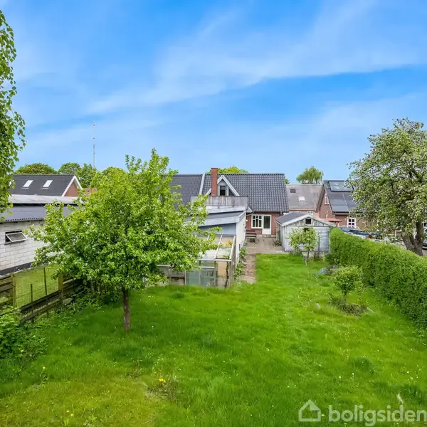Huset ligger i et grønt område, omgivet af træer og græsbane. I baggrunden ses flere huse med skrå tage.