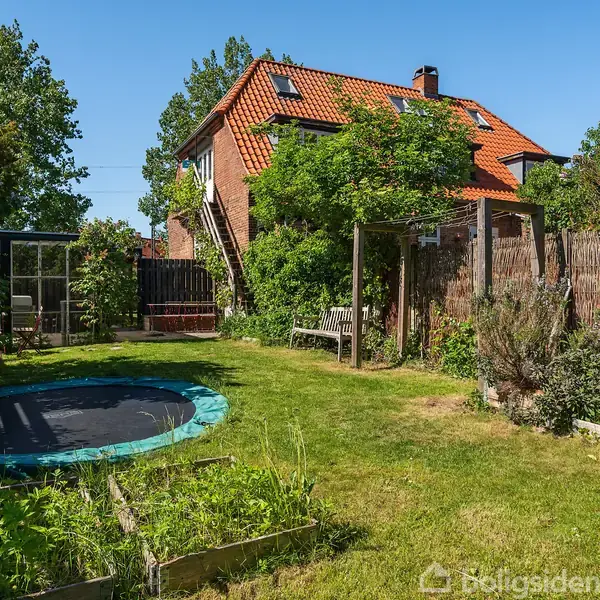 Et rødt murstenshus med rød tegltag er omgivet af en frodig have. Foran huset står en trampolin og en lille plantekasse. En gangsti fører til en bagvedliggende terrasse.