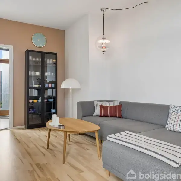 Grå hjørnesofa med puder, placeret i lys stue. Sofabord foran sofa, høj lampe ved siden. Modsat væg er der et vitrineskab. Dør fører til anden del af hjemmet.