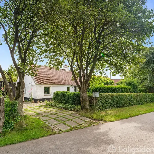 Et hvidt hus med rødt tag står mellem træer. En sti fører til hoveddøren. Grøn hæk omkranser haven tæt på en stille vej. Skraldespande står ved indkørslen.
