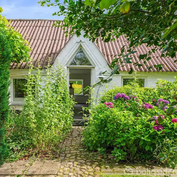 Hus med rødt tegltag, omgivet af grønne buske og blomster, ligger for enden af en brostensbelagt sti. Dør og vinduer er symmetrisk placeret i den hvide facade.