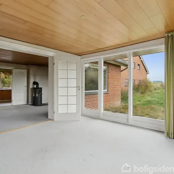 Dobbelt glasdør står åben, leder fra et lyst rum med træloft til en anden stue. Store vinduer giver udsigt til en græsplæne og et rødt murstenshus. Gardiner hænger til højre.