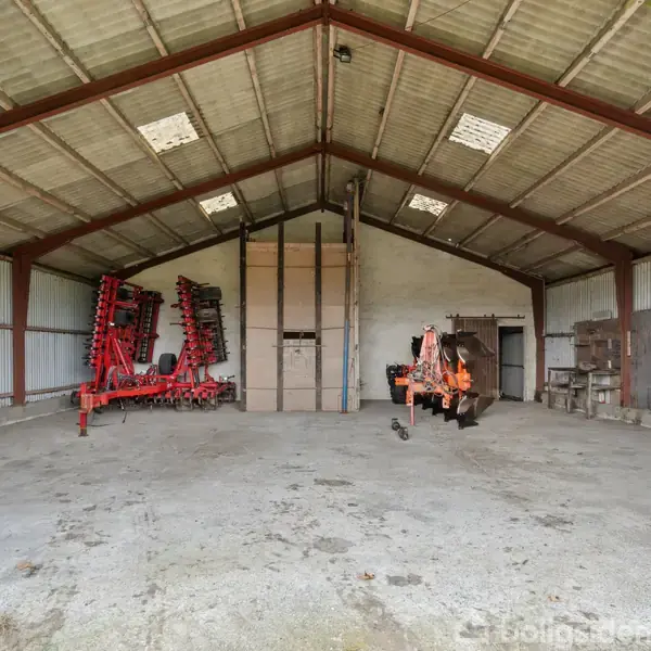 En tom lade indeholder landbrugsmaskiner, herunder en stor rød plov og en orange maskine, placeret på en betonbund. Væggene og taget er af metal, med åbne porte.