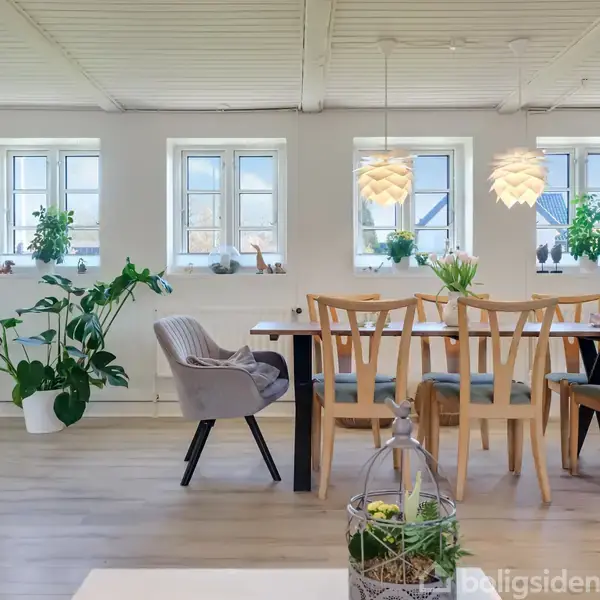 Spisebord med seks stole står under to hængende lamper. Store vinduer i baggrunden med planter på karmen. Lys, rummelig stue med lyse trægulve og hvide vægge.