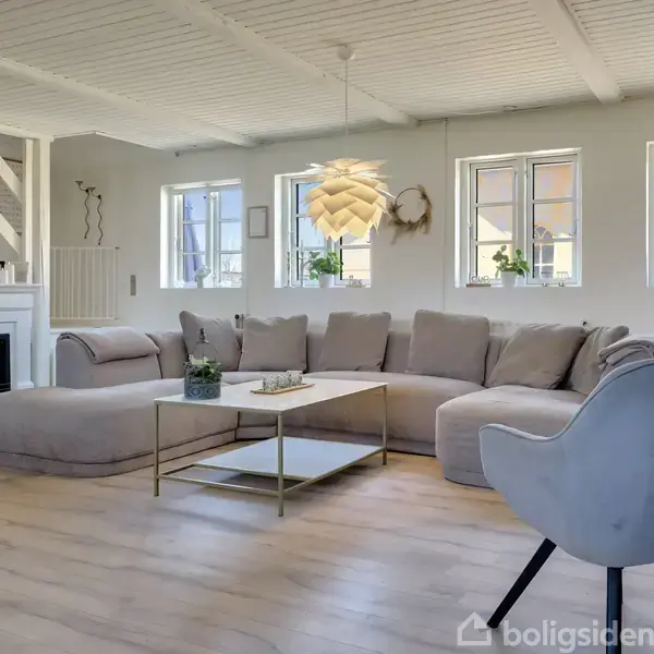 En beige hjørnesofa står i en lys stue med trægulv, ved siden af en hvid pejs. En moderne, hvid loftlampe hænger over sofabordet. Tre vinduer lader naturligt lys ind.