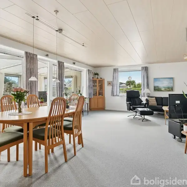 Et spisebord med seks stole står centralt i en lys stue. Bordet har blomster og hænger under moderne lamper. Rummet har store vinduer, lyse vægge og loungemøbel i hjørnet.