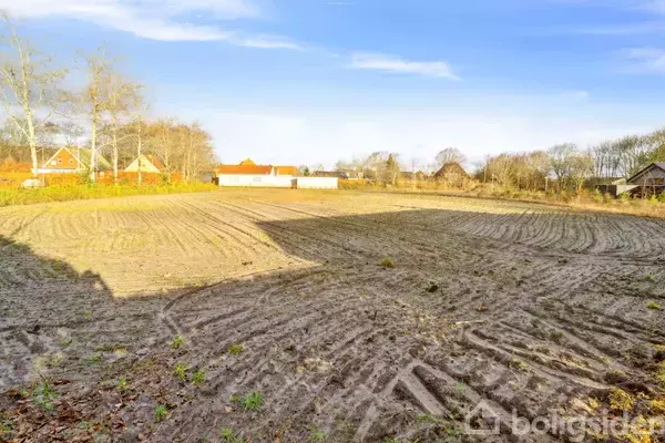 Åbent, jævnt pløjet jordstykke ligger stille i et landligt område omgrænset af nogle spredte huse og træer under en klar blå himmel.