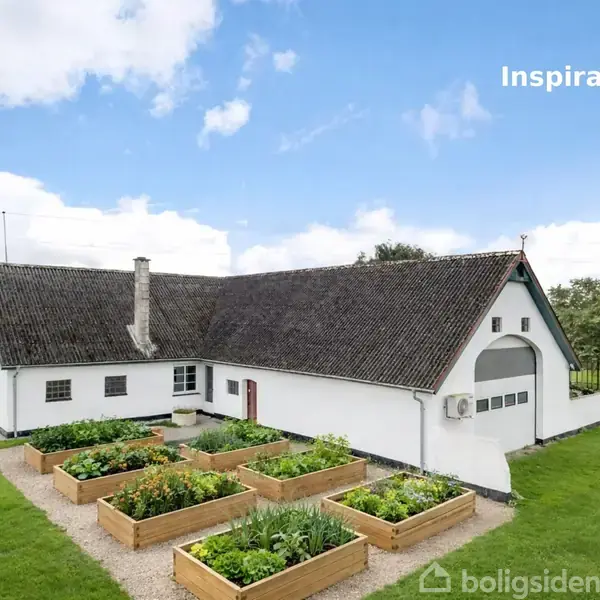 Hvidt hus med traditionelt sadeltag omgivet af grøn have. Foran huset ligger seks højbede med grøntsager. Blå himmel med skyer i baggrunden. Teksten "Inspiration" ses øverst.