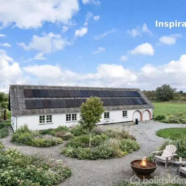 En hvid bygning med solpaneler på taget står i en gårdhave. To stole og et bålsted er foran bygningen. Grønt landskab omkranser området. Tekst: "Inspiration".