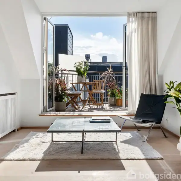 En stue med glasbord står på et hvidt tæppe. Dør åbner ud til balkon med træstole og planter. Hvide vægge, indrammede billeder og lysindfald giver rummet en lys atmosfære.