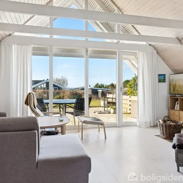 En stue med en grå sofa og en hvælvet træloft. Store vinduer åbner mod en terrasse med bord og stole. En brændeovn står i hjørnet.