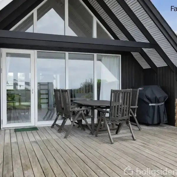 Udendørs terrasse med mørkt træbord og stole foran store glasdøre under en sort trætaget gavl. Gasgrill til højre og "Facadeforslag" skrevet øverst i billedet.