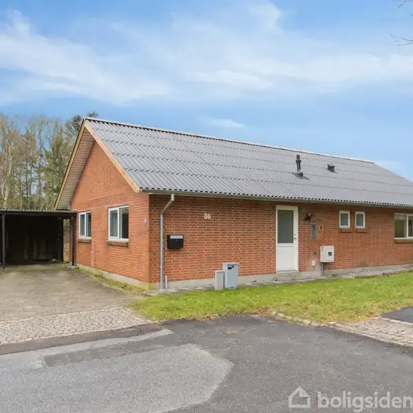 Murstensvilla med gråt tag står stille ved siden af en carport og indkørsel. Græsplæne omgiver huset, beliggende i et villaområde med træer i baggrunden.