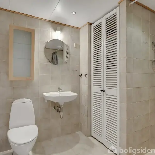 Et badeværelse med en hvid håndvask monteret på væggen under et rundt spejl, toilettet står til venstre. Flisevæggene skaber en ren, lys atmosfære.