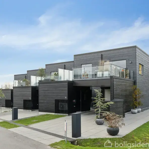 Et moderne, sort træhus står stille i en forstadsmiljø med fladt tag og store vinduer. Foran huset er nogle planter i krukker og en flisebelagt indkørsel langs en vej.