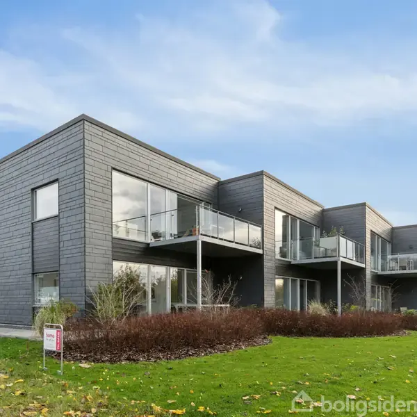 Moderne lejlighedsbygning med store vinduer og altaner omgivet af græsplæner og buske under en blå himmel. En salgsside er synlig i forgrunden.