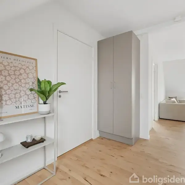Billedramme på en væg viser "Papier Découpé" med blomstermotiv. Konsolbord med plante og lys. Trægulv i lyst rum leder til stue med sofa og store vinduer med udsigt.