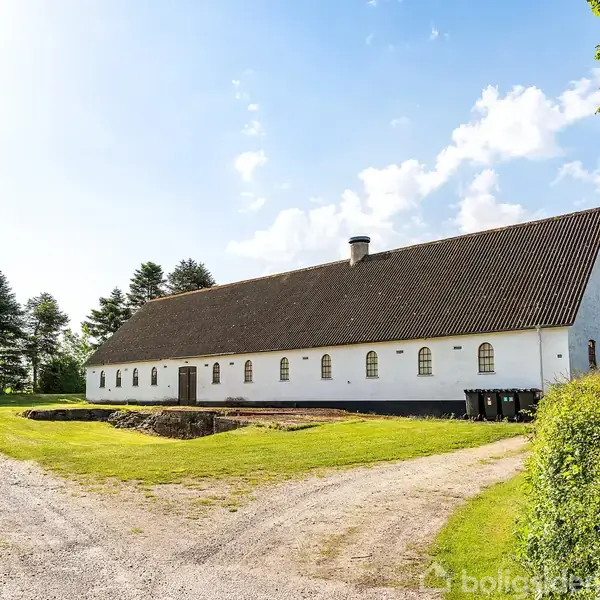 En lang, hvid bygning med brunt tag står stille. Omkring bygningen er der en grusvej, grønne træer og en græsplæne under en blå himmel med skyer.
