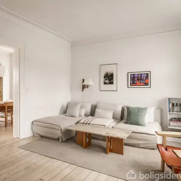 Sofa med puder står i en stue ved siden af et lille bord og en stol. På væggen over sofaen hænger to billeder. I baggrunden ses en spisestue med et bord og stole.