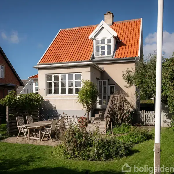 Et lille hus med rødt tegltag står i en have. Der er et bord og stole på en fliseterrasse foran huset, omgivet af grønne buske og græsplæne.