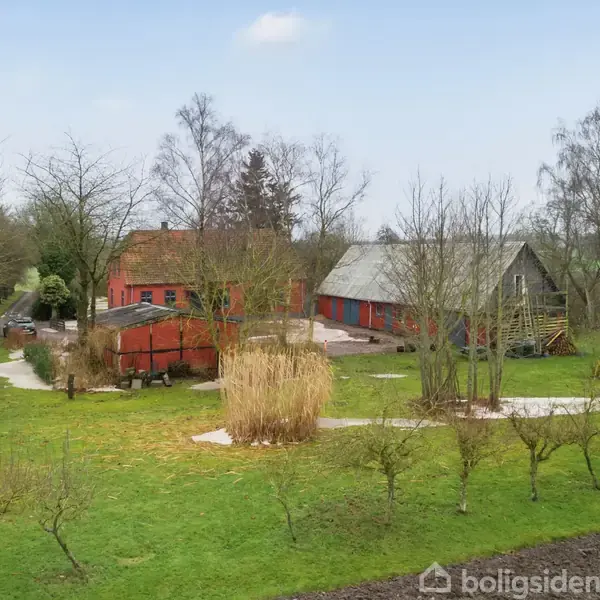 Røde landbrugsbygninger ligger omgivet af grønne marker og træer. En grusvej fører frem til gården, som er omkranset af en sparsom beplantning af træer, der står uden blade.