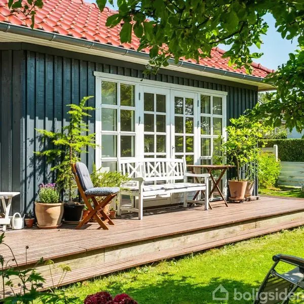 Husets terrasse har hvide møbler og planter i krukker. En stol står ved siden af et bord. Omgivet af en grøn have og træer, med sollys.