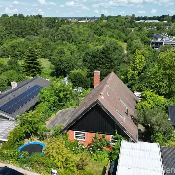 Et hus med skråt tag står i en frodig, grøn have. Solpaneler er synlige på naboens garage. Omgivet af tæt skov og flere huse.