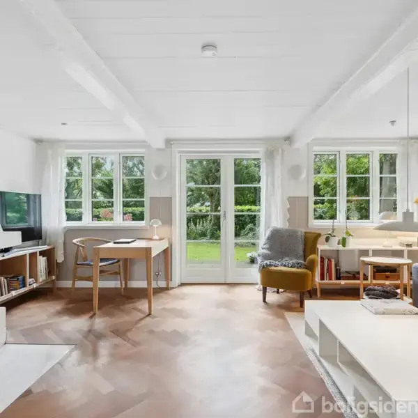 Stue med sofa og bord, i en lys indretning. Store vinduer og døre med udsigt til en grøn have. Hylde med planter, lænestol ved lampe, skrivebord ved TV.