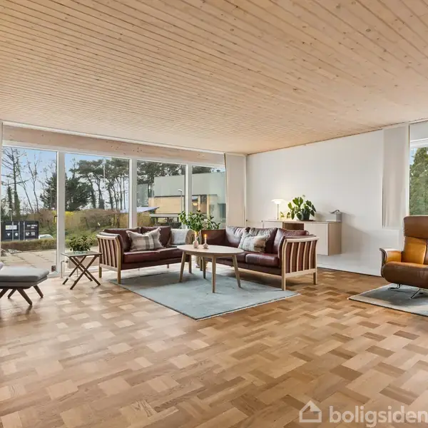 Stue med moderne møbler, herunder sofaer og lænestole, arrangeret på tæpper. Store vinduer giver udsigt til en have. Trægulv og loft skaber en varm atmosfære.