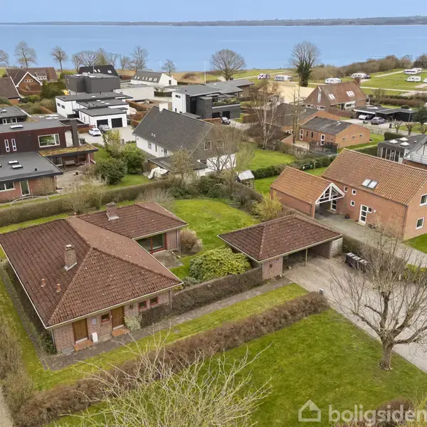 Boliger med røde tage står stille i en grøn have. Omkring dem er der flere huse i et villakvarter med udsigt til en fjord i baggrunden og nogle marker.