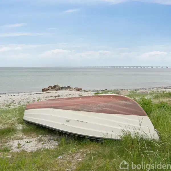 En omvendt båd hviler på en sandet strand, omgivet af græs. I baggrunden ses havet og en lang bro under en blå himmel med lette skyer.